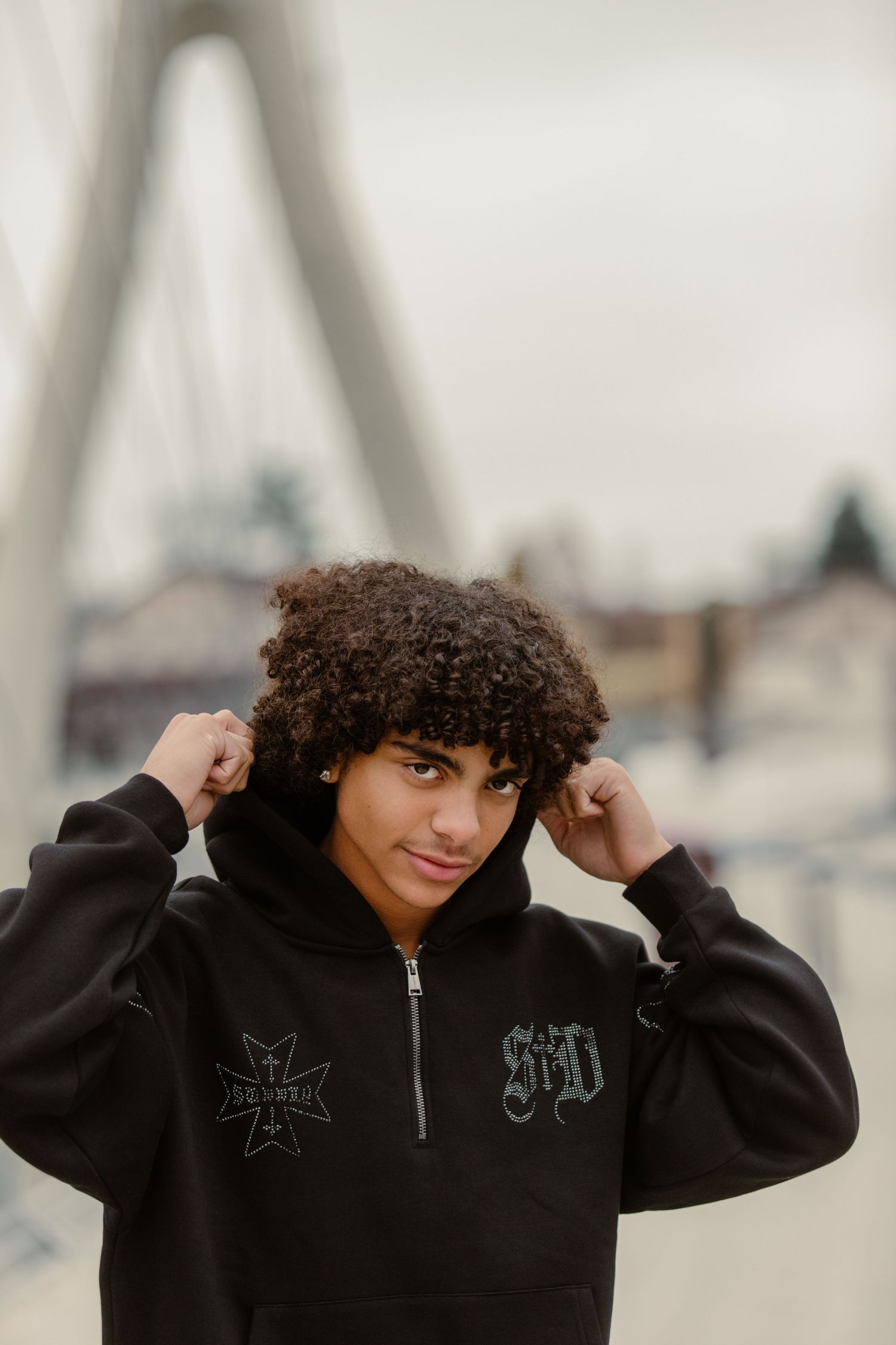 Teenage Senior guy stands in front of bridge in Dublin, Ohio, pulling his hood up for dramatic Senior portrait photo