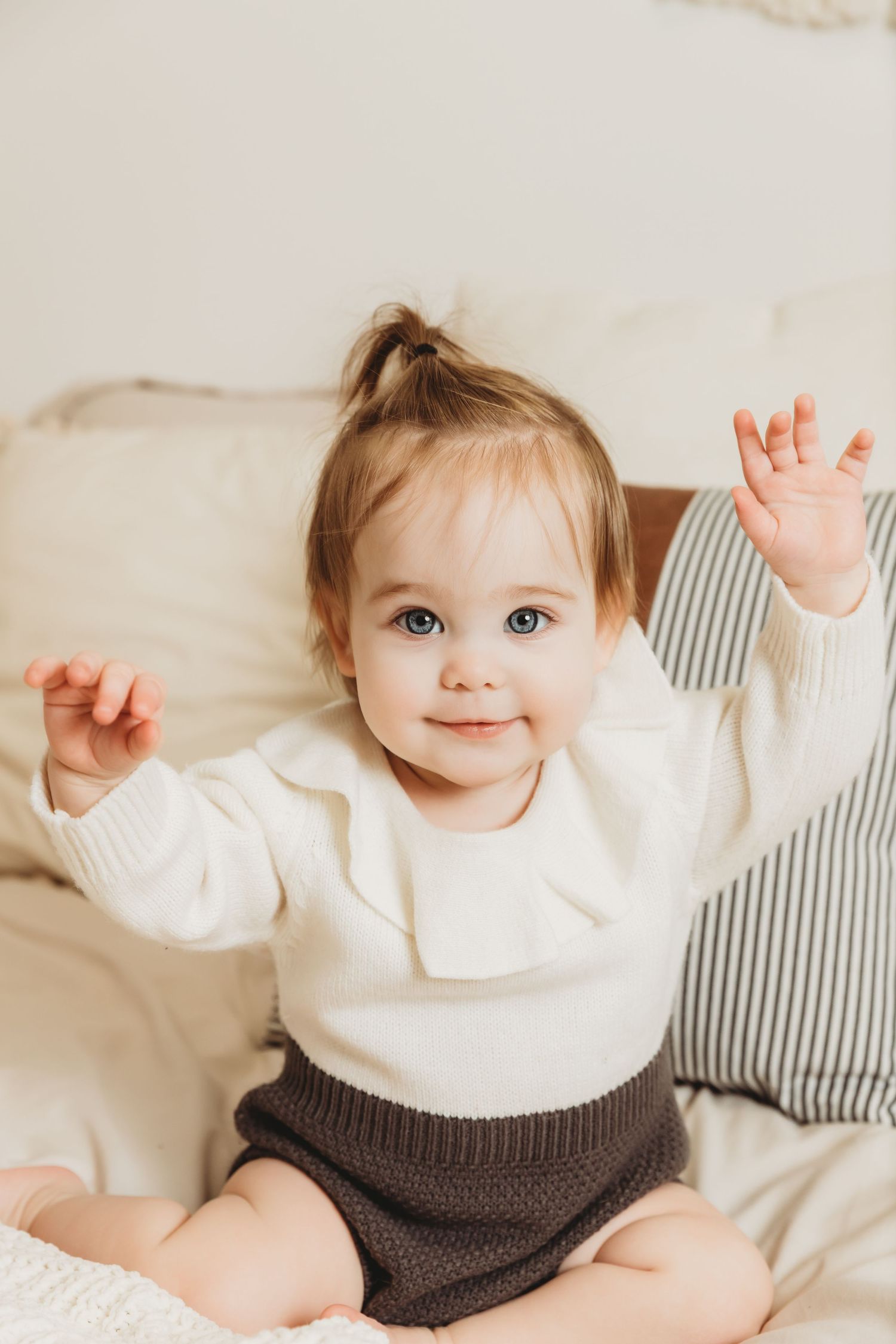 Baby girl sitting on bed smiling with hands raised for 1st birthday baby milestone photos with Tami Harris Photography in-studio