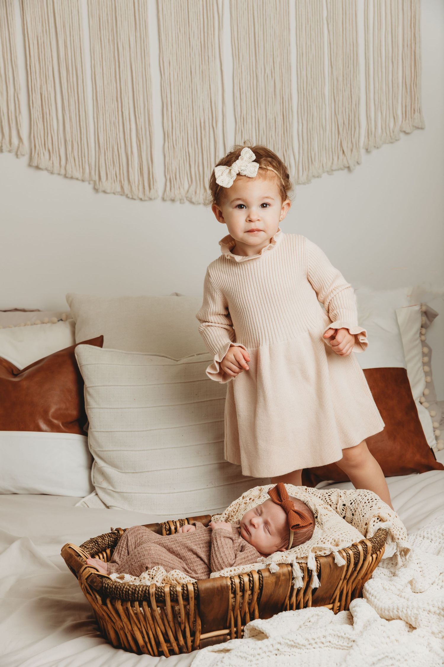 2-year-old big sister stands on bed next to her baby sister in a basket during newborn photoshoot with Tami Harris Photography