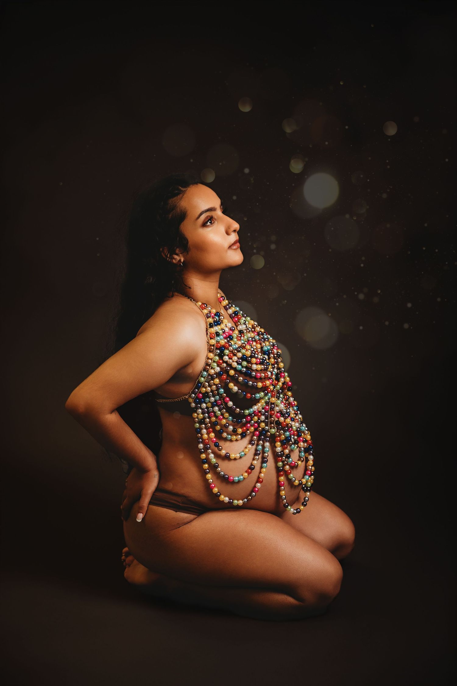 Pregnant mother dressed in beaded necklace kneels on studio backdrop while looking up for Maternity photoshoot with Tami Harris Photography