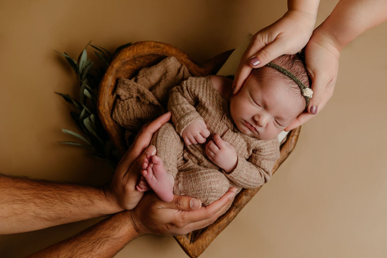 parents support newborn baby girl in hands while she sleeps in a wooden heart-shaped bowl during a photo session w/ Tami Harris Photography