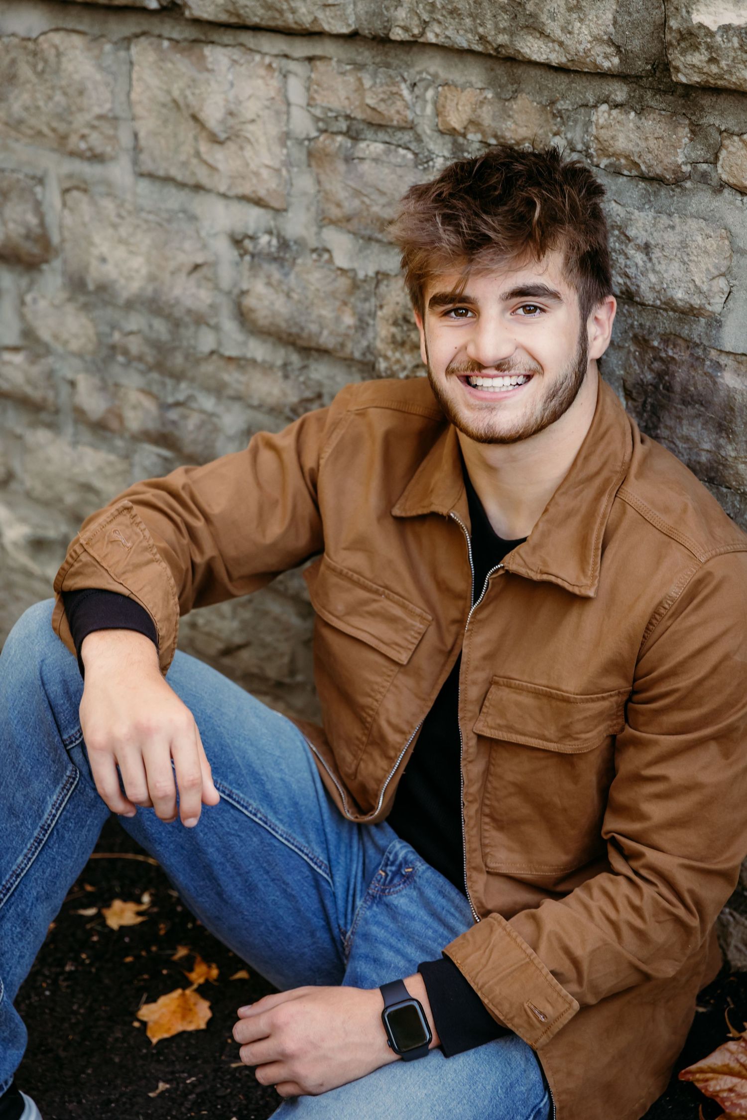 Guy smiles sitting casually against stone wall at Inniswood in Westerville, Oh for Senior portrait photoshoot with Tami Harris Photography