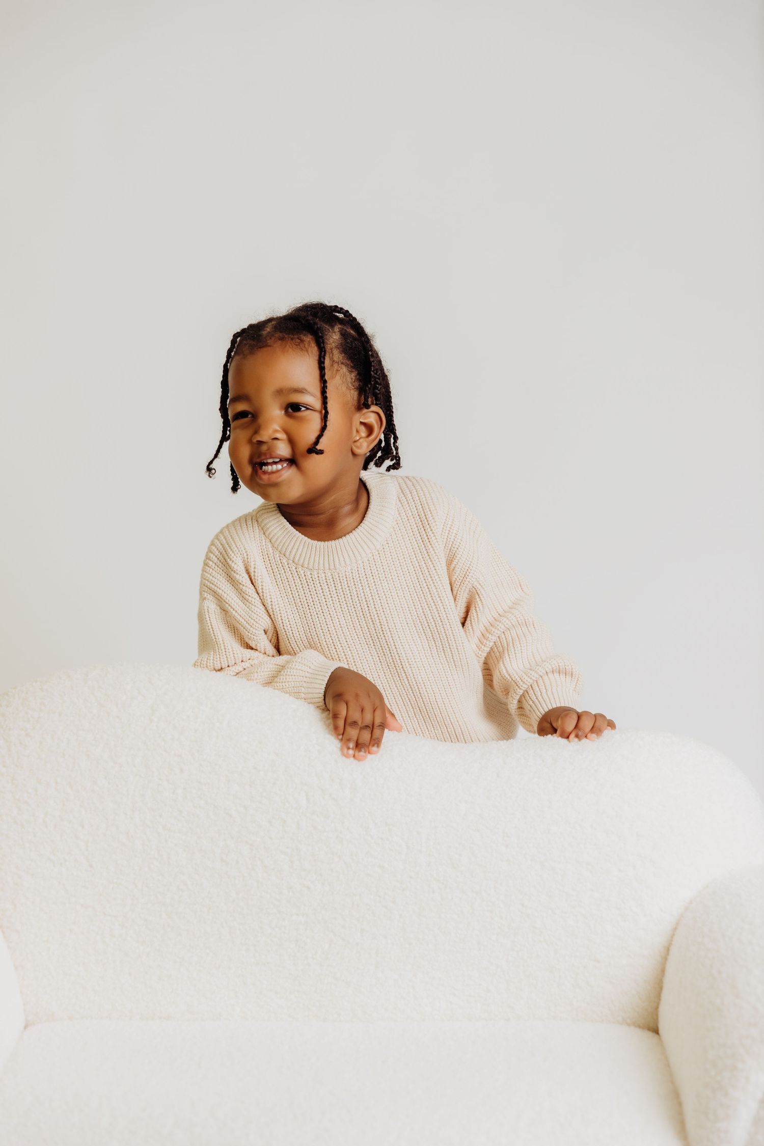 Smiling boy with braids stands behind child-size couch in studio milestone session for 2nd birthday photoshoot with Tami Harris Photography