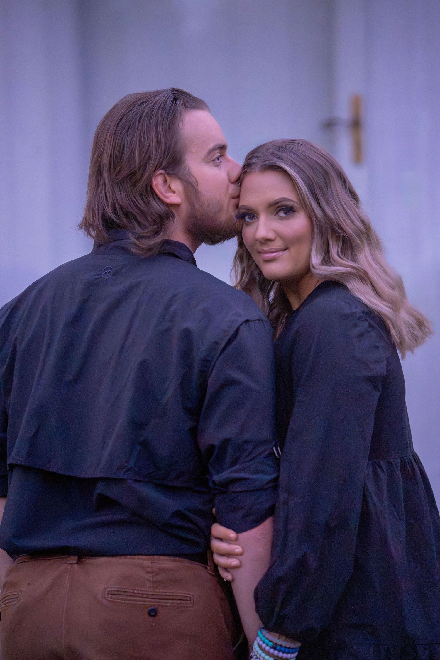 Couple in matching dark outfits sharing romantic moments against purple backdrop.