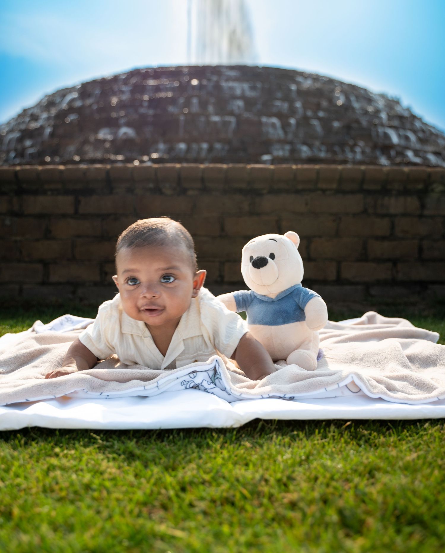 Baby photoshoot on grass with white blanket and teddy bear props.