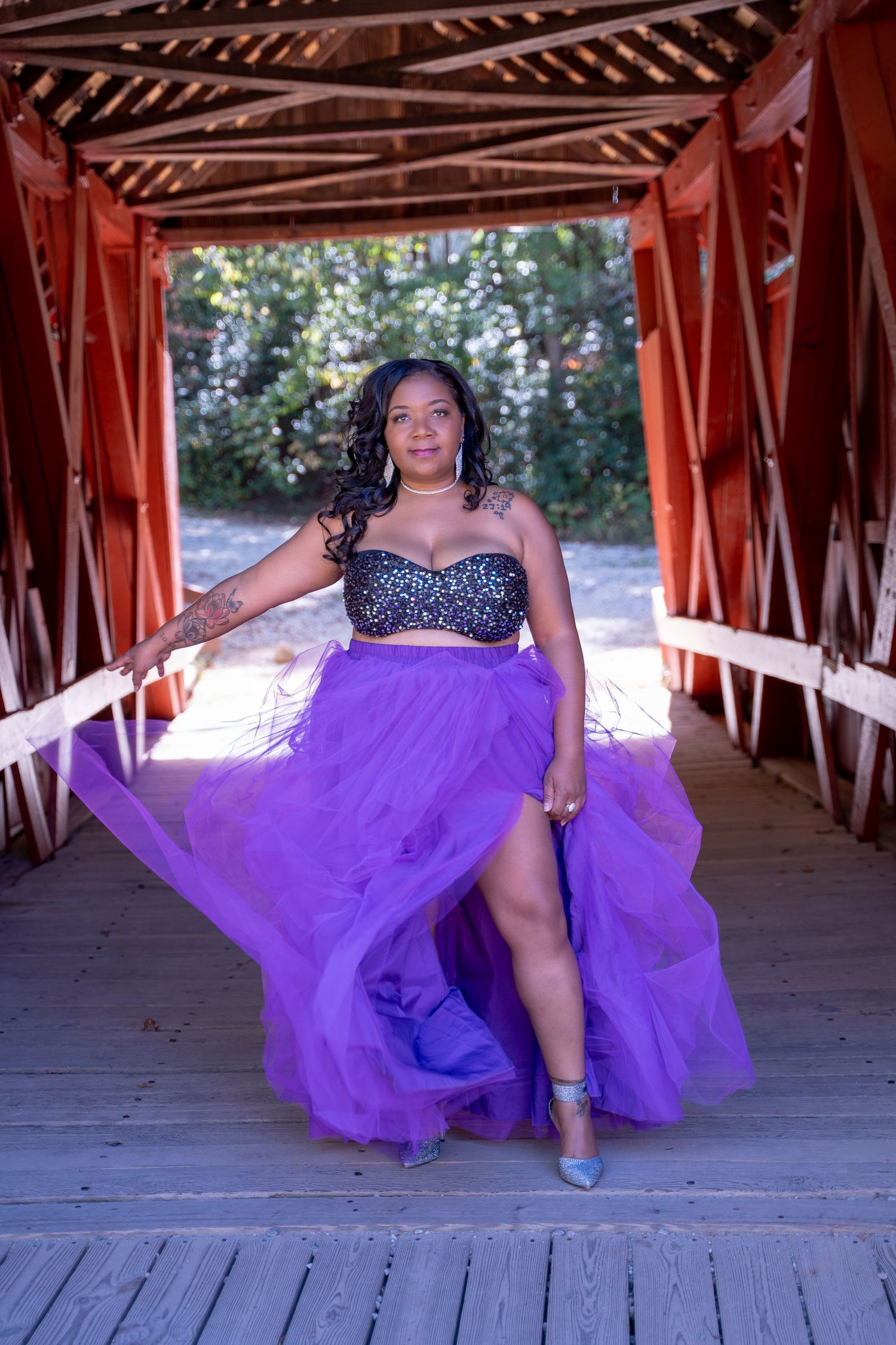 Person in purple ball gown poses dramatically in covered bridge with natural lighting.