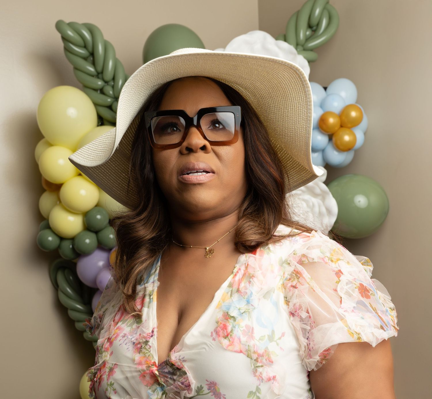 Woman in floral dress and sun hat poses against pastel balloon display.