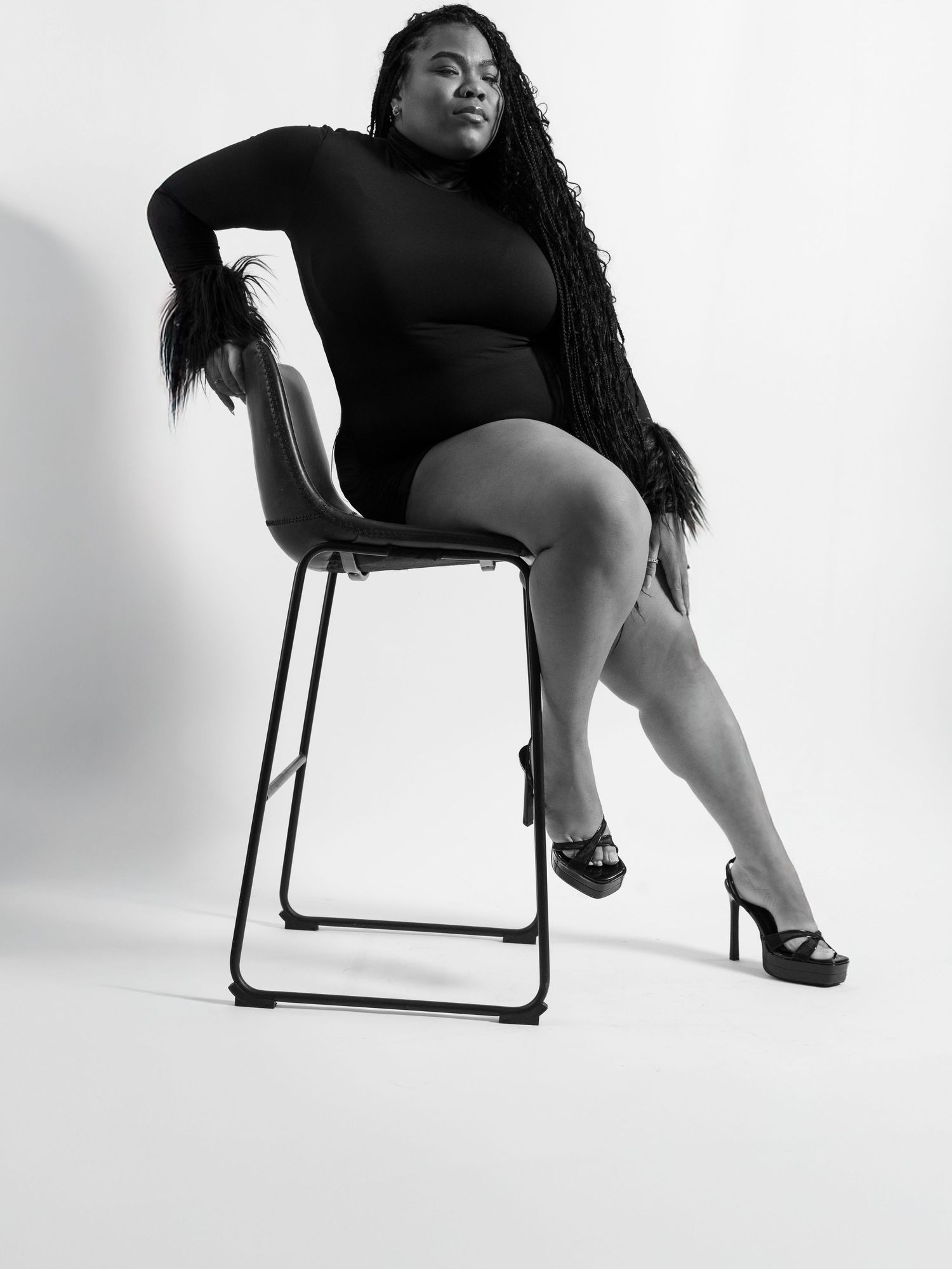 Elegant black and white portrait of someone in dark attire posing confidently on a bar stool.