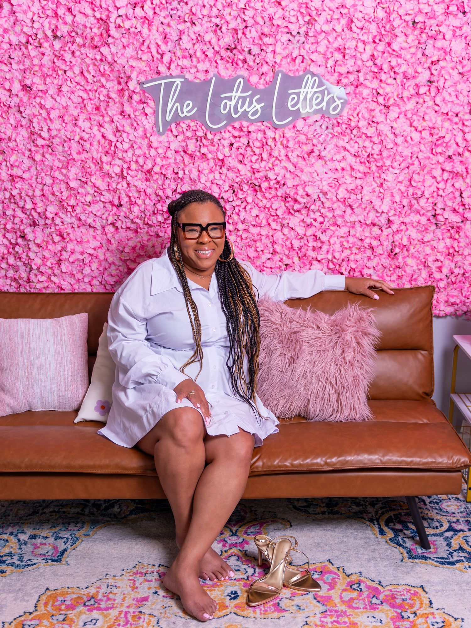 Relaxed portrait against pink floral wall with 'The Lotus Letes' neon sign and brown leather couch.