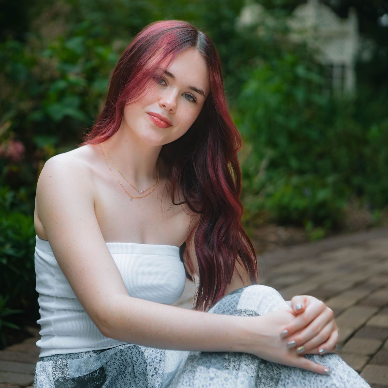 Portrait of someone with long red hair wearing white strapless top sitting outdoors with greenery in background in Youngstown, Ohio.