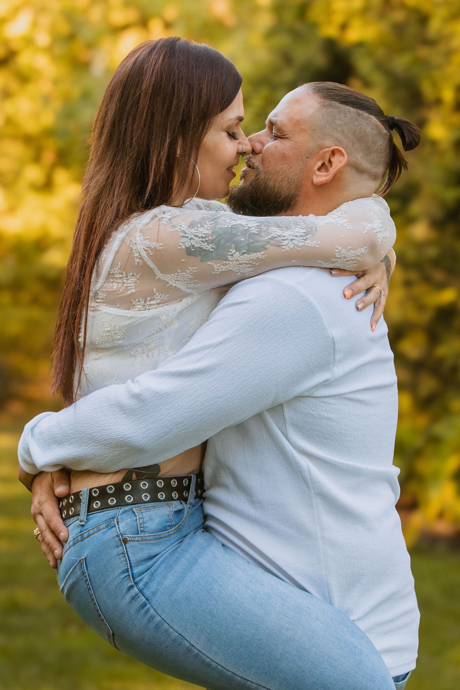 Couple in jeans and white embracing outdoors with golden autumn foliage in background in Youngstown, Ohio.