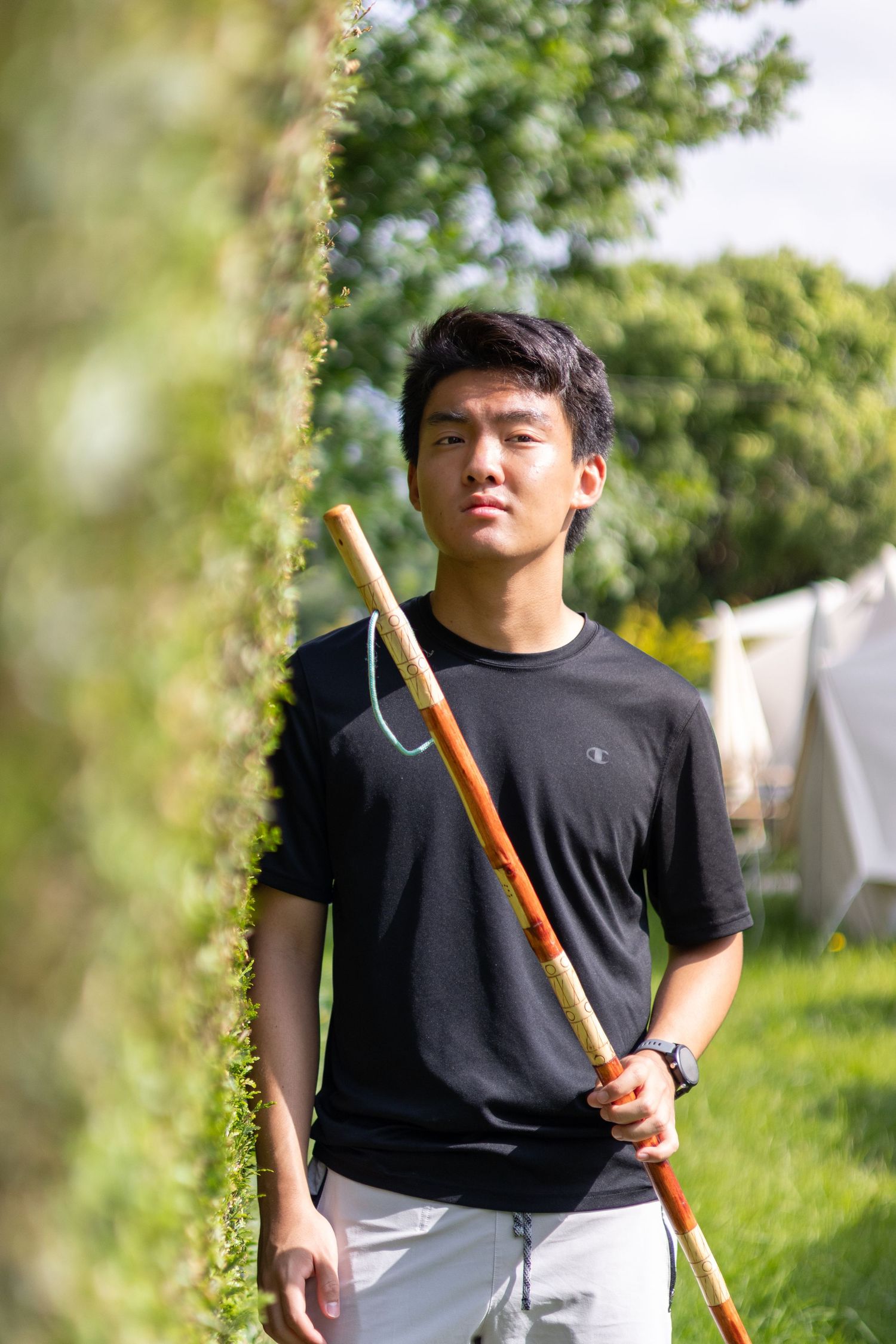 A person in a black t-shirt holds a long wooden pole while standing outdoors in a green garden setting. Luke Ye photography, Luke Ye,