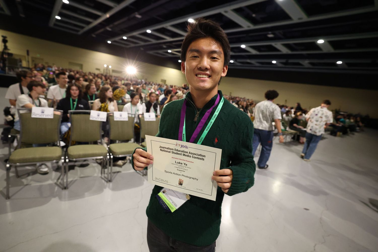 Student in green sweater proudly displays academic certificate at awards ceremony in large auditorium. Luke Ye photography, Luke Ye,