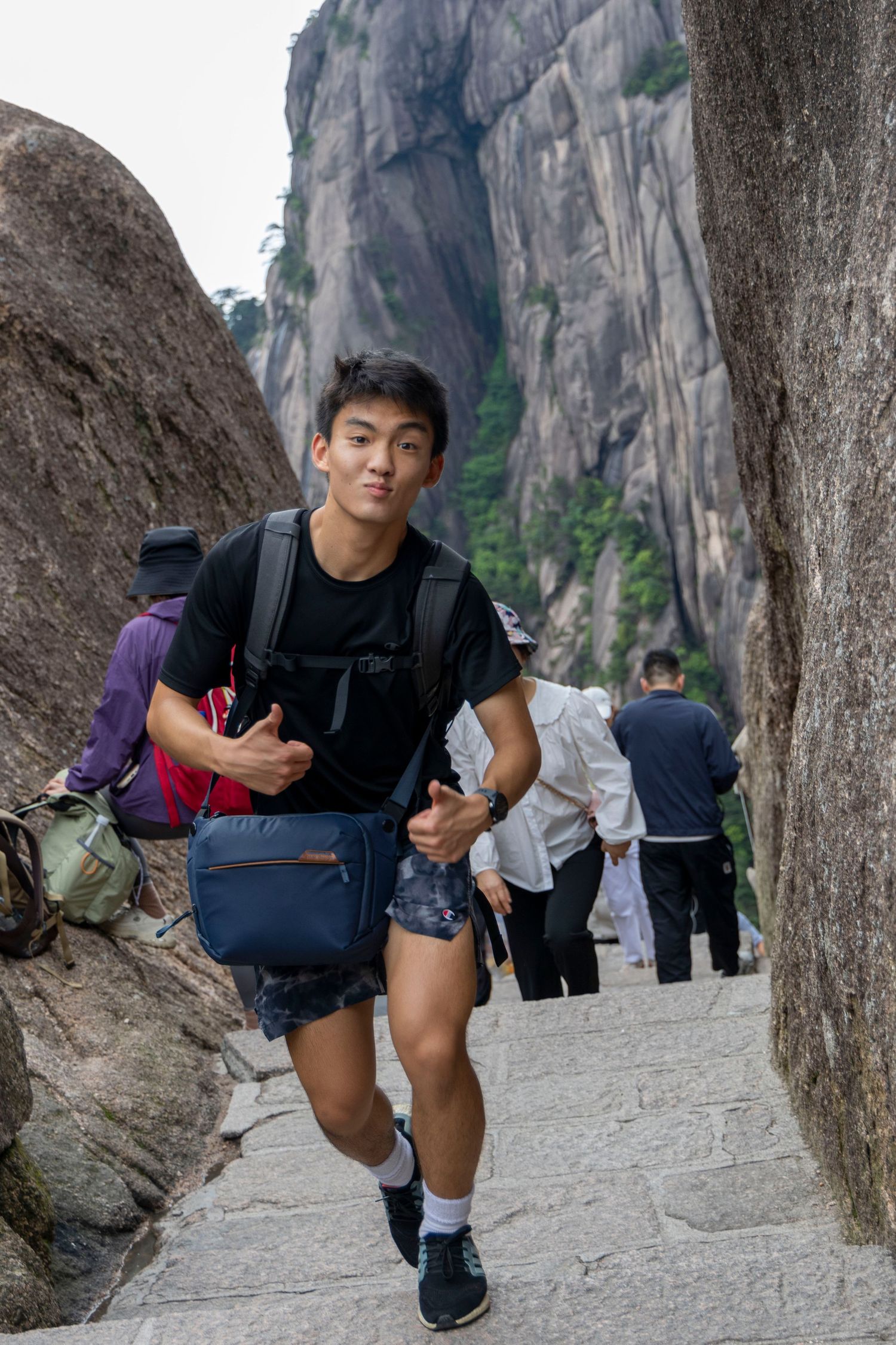 A person poses playfully between rock formations on a mountain hiking path with other hikers visible in the background. Luke Ye photography,