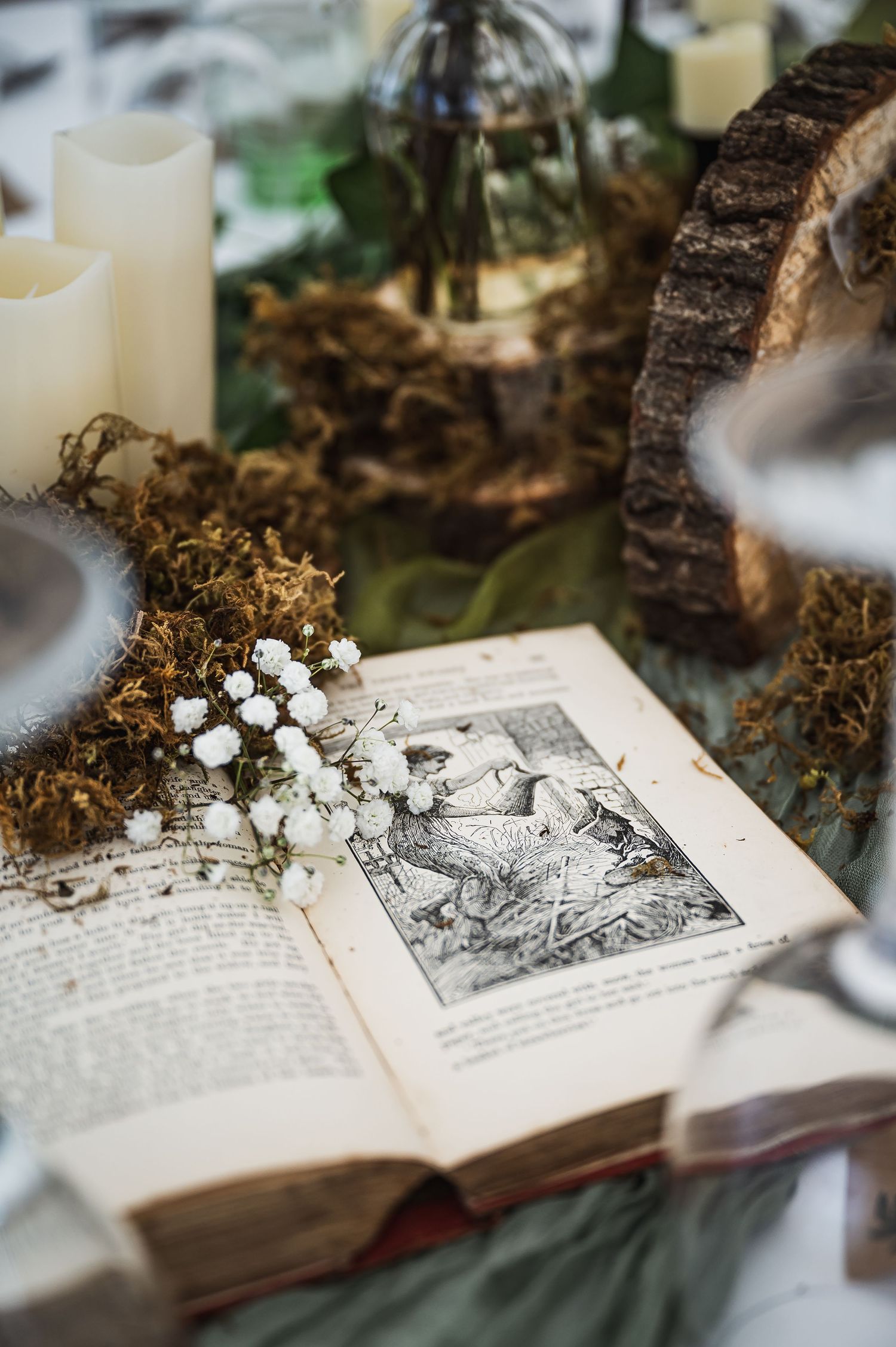 Rustic wedding table decor with open vintage book, white gypsophila flowers, moss, candles, and woodland elements.