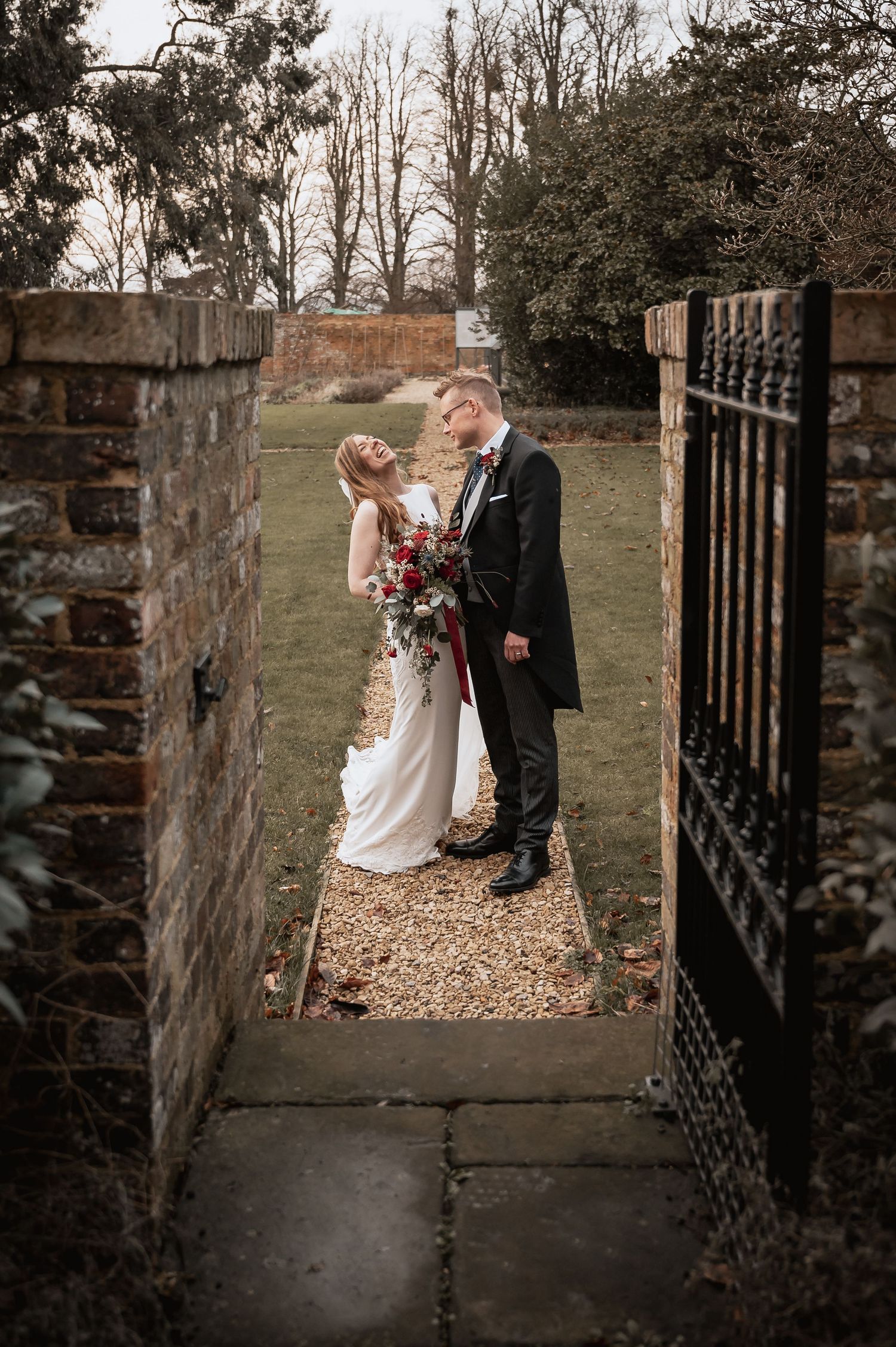 Bride and groom share a tender moment framed by a rustic stone gate, bride holding red flowers.