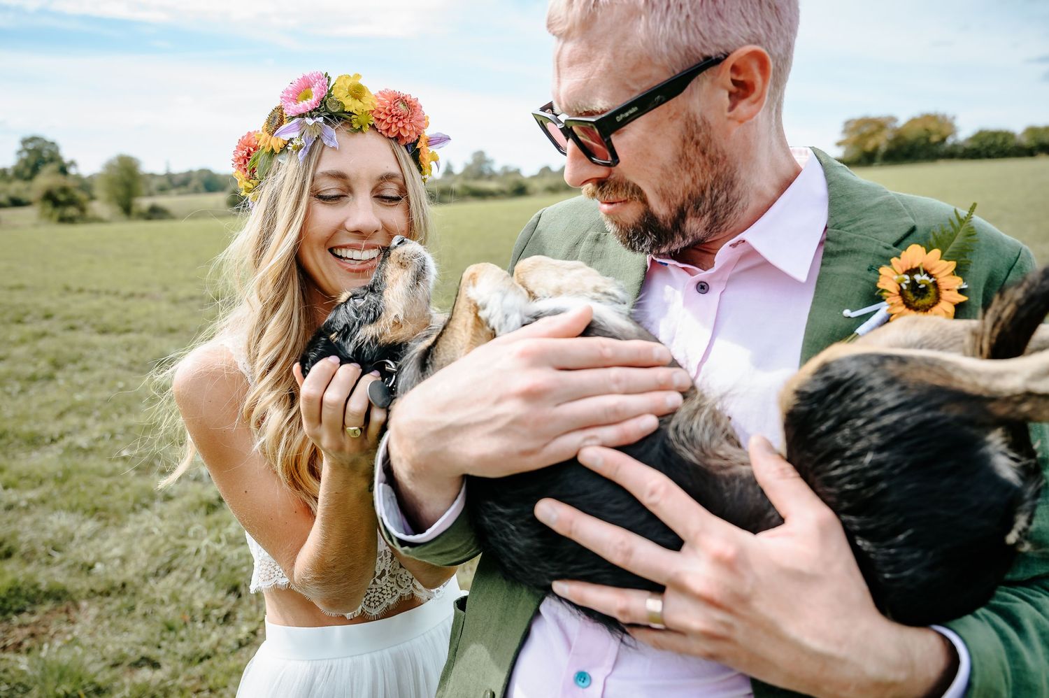 Bohemian bride and groom holding small animals in a sunny field with floral accessories.