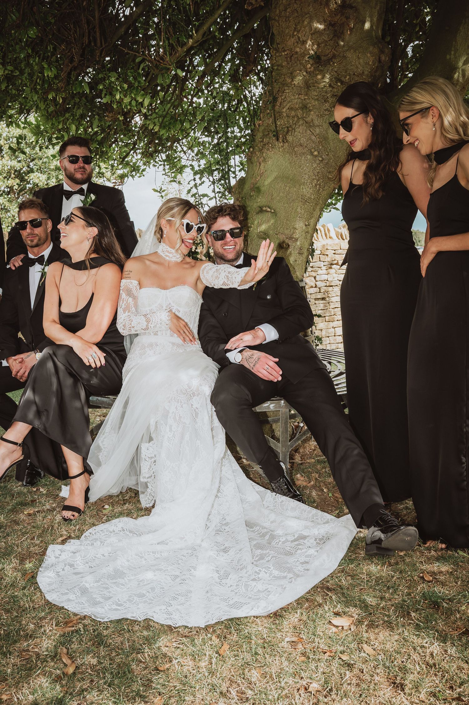 Bride and groom wearing sunglasses celebrate with bridesmaids and groomsmen in black attire outdoors.