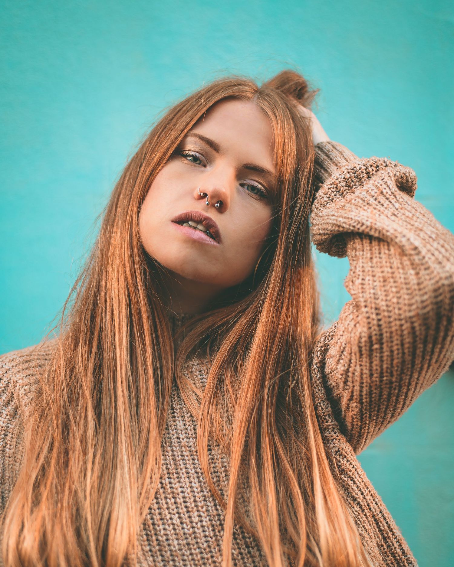 Young woman with long auburn hair and nose ring in beige sweater against turquoise wall.