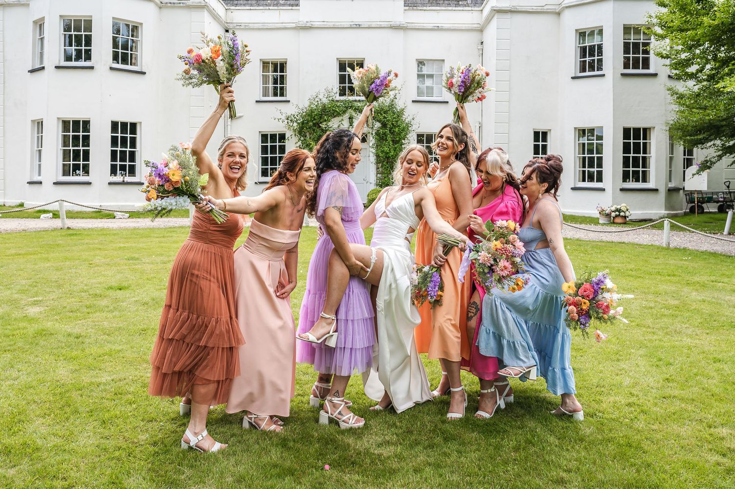 Bride and colorful bridesmaids in mixed dresses celebrating with bouquets outside a white manor house.