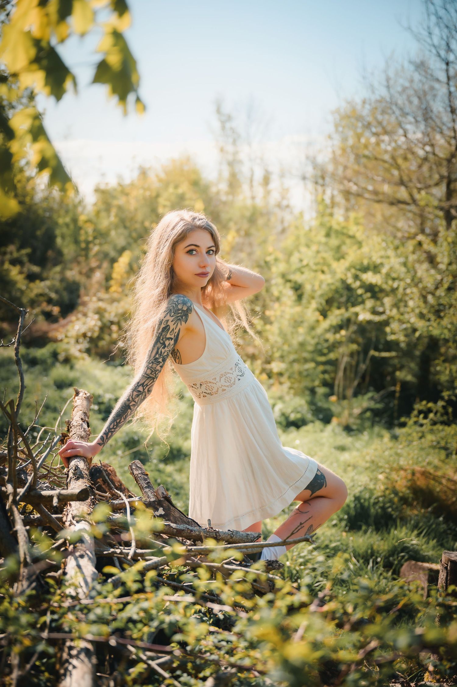 Blonde woman with tattoos in white dress posing in a sunny green forest setting.