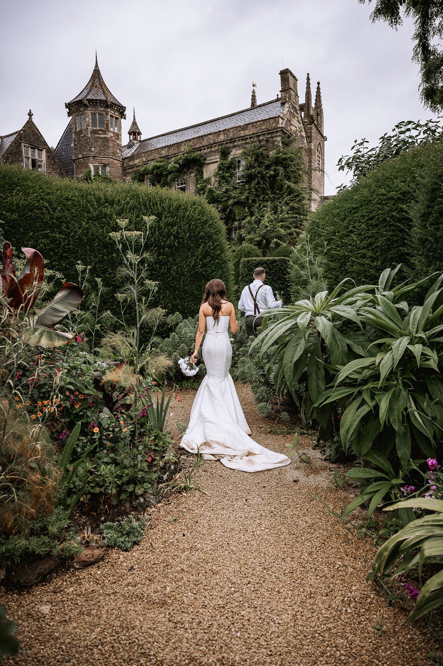Bride and groom walking along a garden path toward a grand Gothic manor on their wedding day.