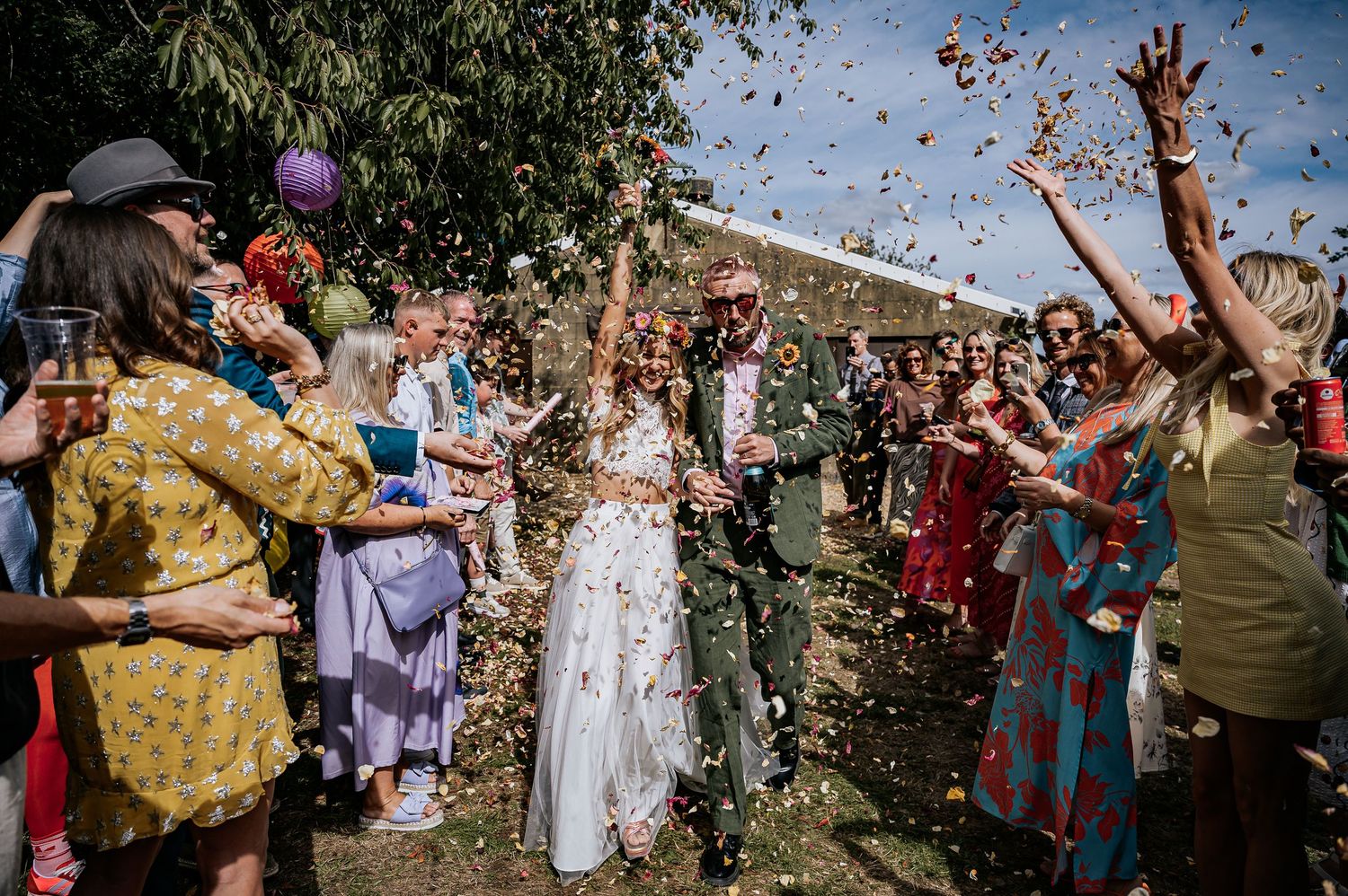 Bride and groom walk through colorful confetti shower surrounded by cheering wedding guests outdoors.