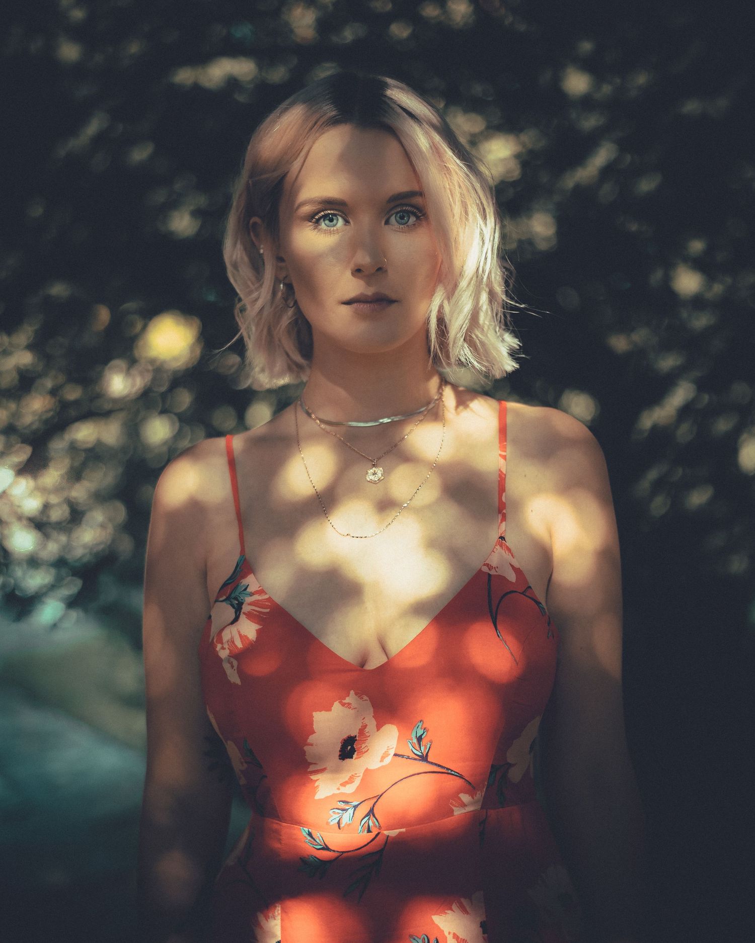 Blonde woman in red floral sundress stands in dappled sunlight with trees in the background.
