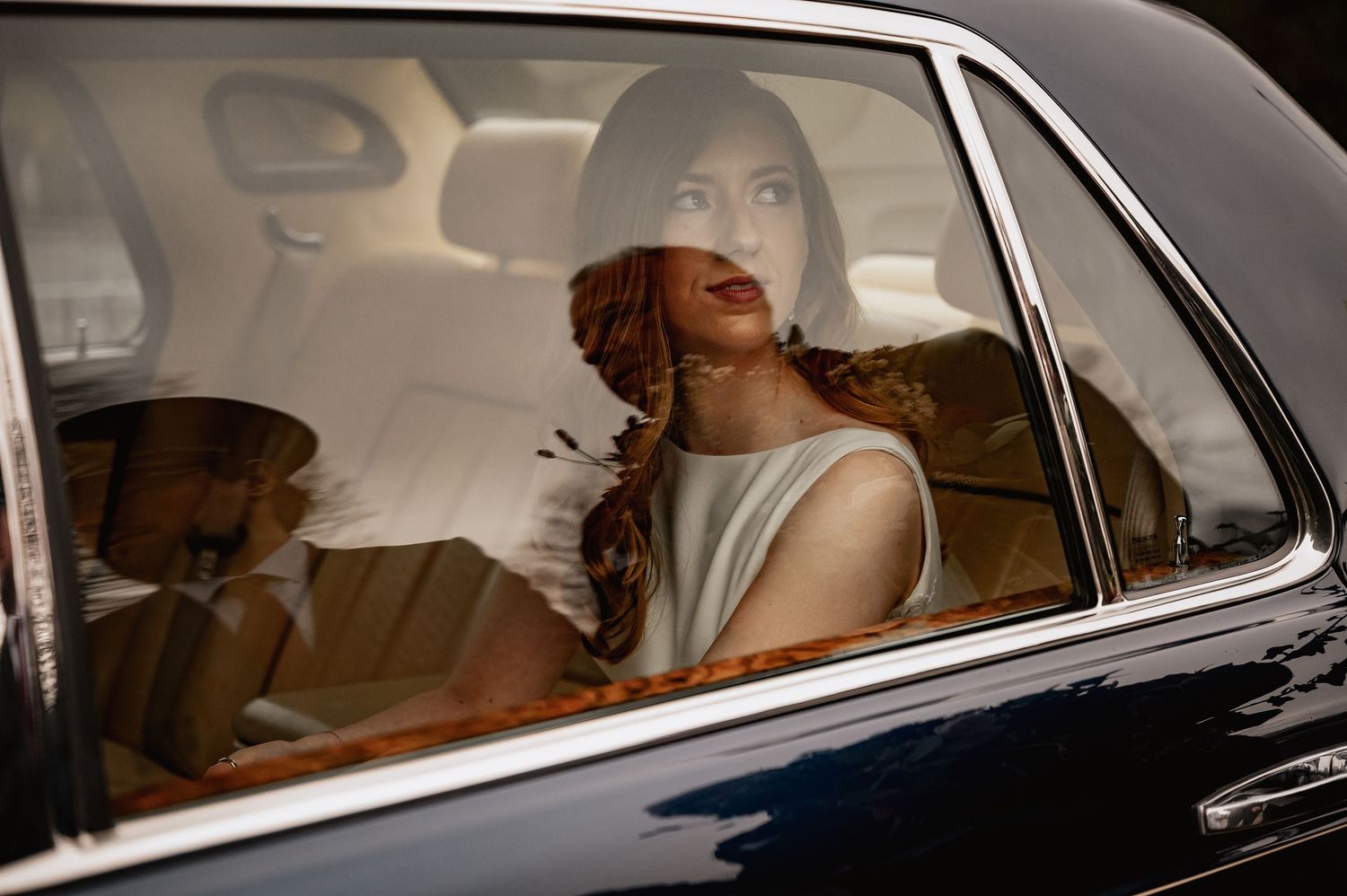 Elegant bride with red hair sits in a vintage car, wearing a white dress and feathered hair accessory.