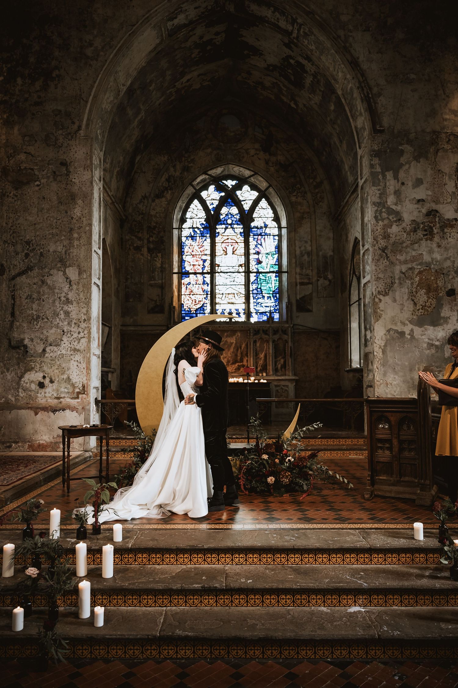 Bride and groom sharing first kiss at gothic church altar with crescent moon backdrop and stained glass window.