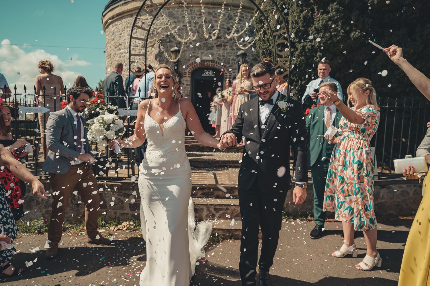 Bride and groom exit ceremony through confetti shower, guests celebrating outside a stone building on a sunny day.