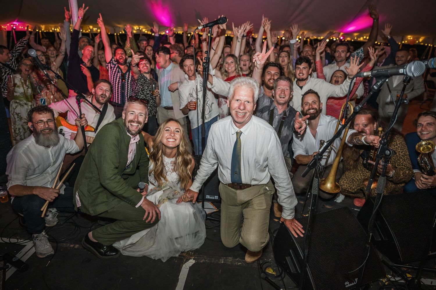 Bride, groom, and guests pose energetically with live band inside a packed wedding reception tent.