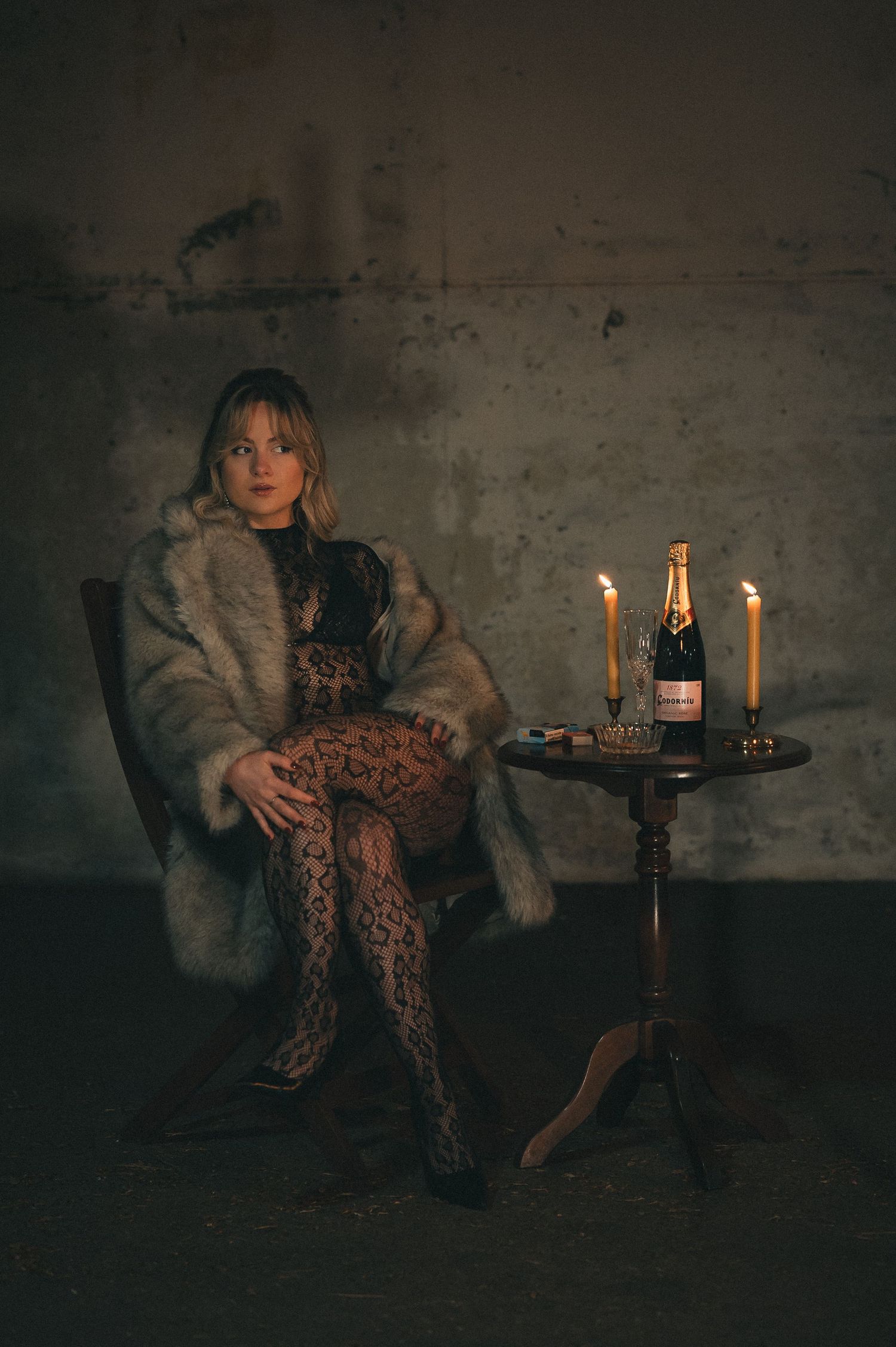 Elegant woman in fur coat and lace dress seated by candlelit table with champagne bottle.
