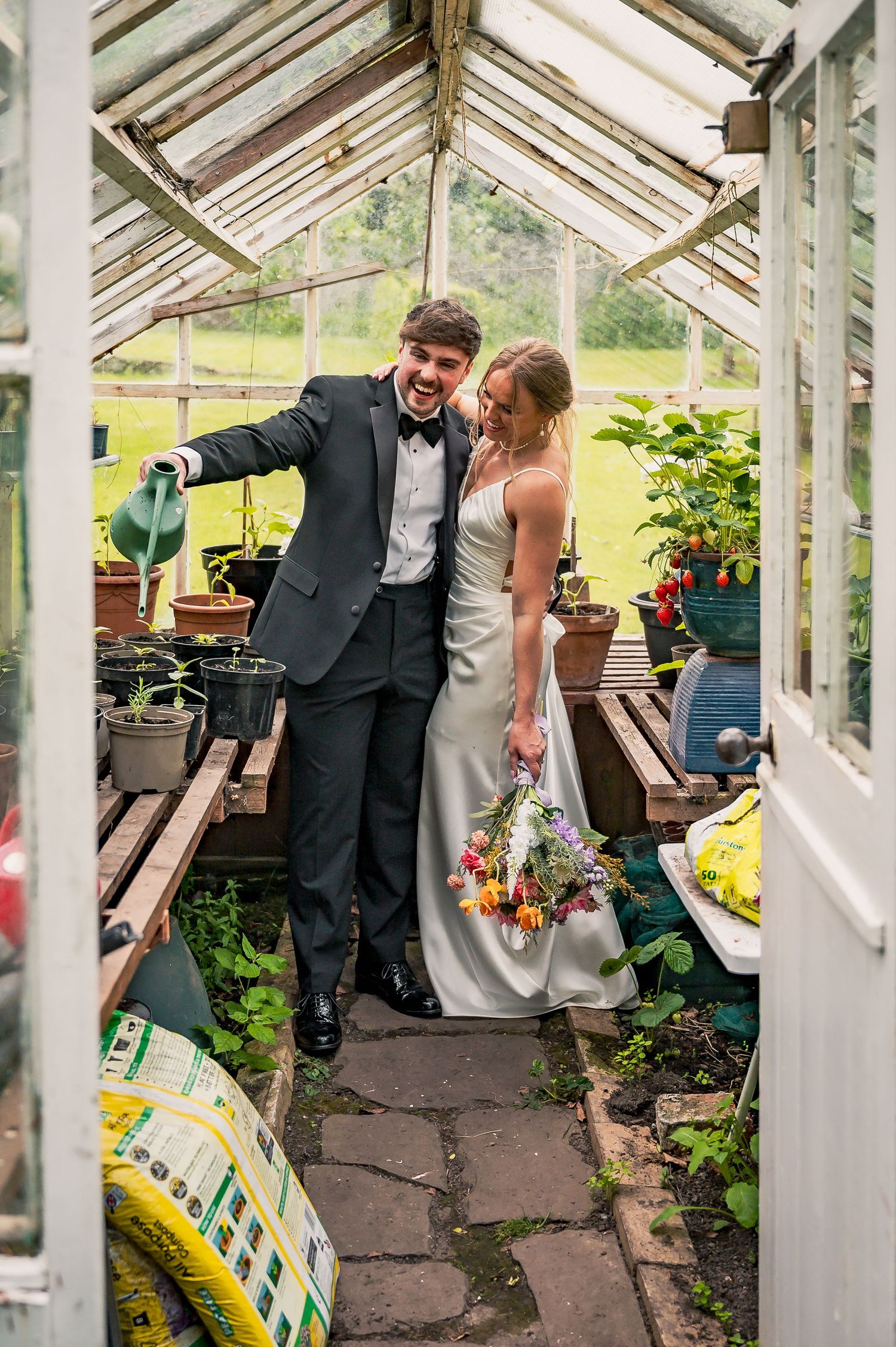 Joyful newlyweds posing playfully inside a rustic greenhouse with potted plants and wildflower bouquet.