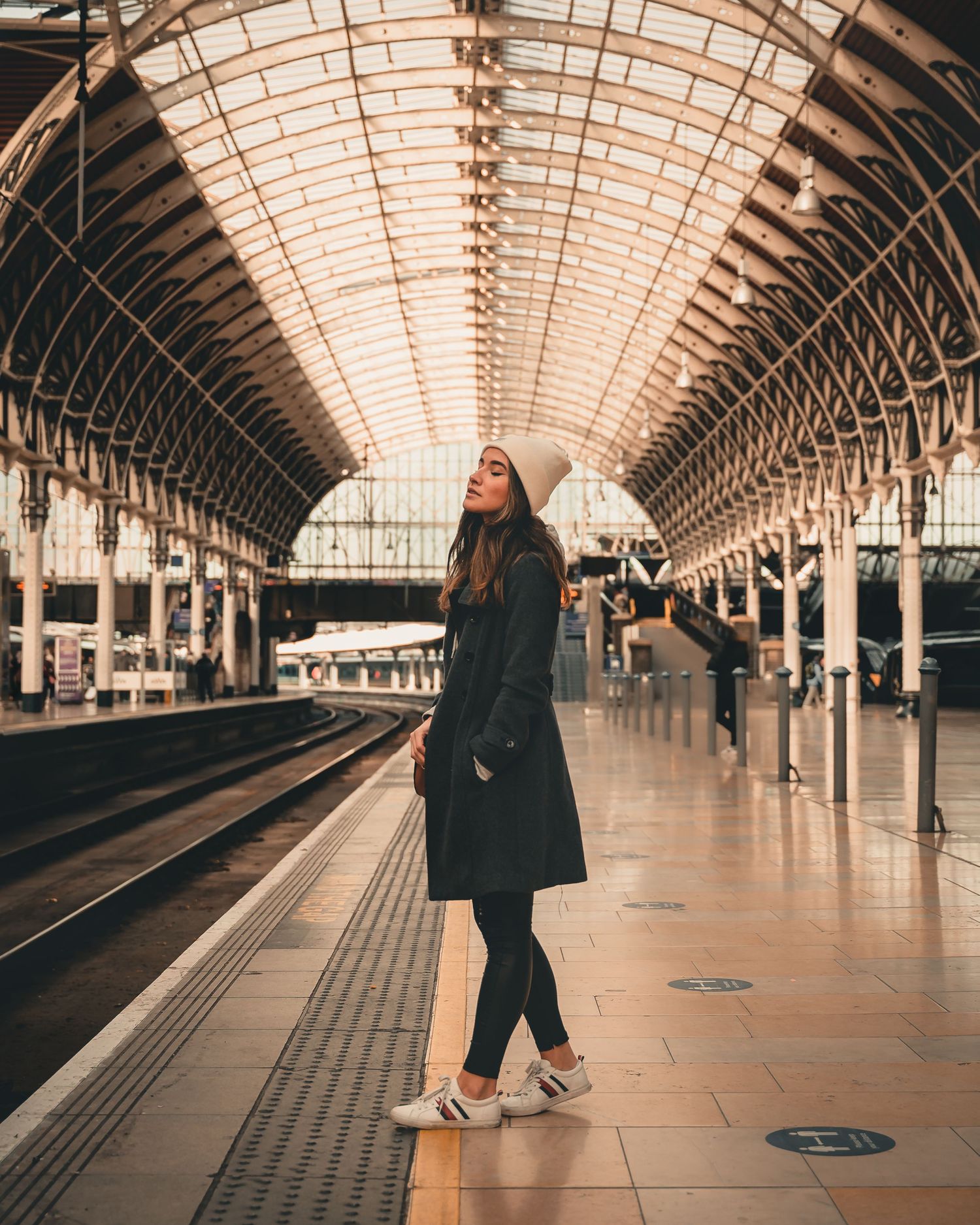Stylish woman in black coat and white beanie posing on platform at grand train station with arched roof.
