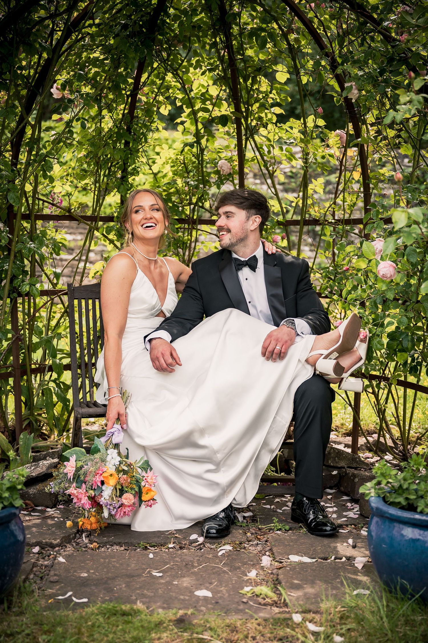 Laughing bride sitting on grooms lap under a garden arbor with pink roses and lush greenery.