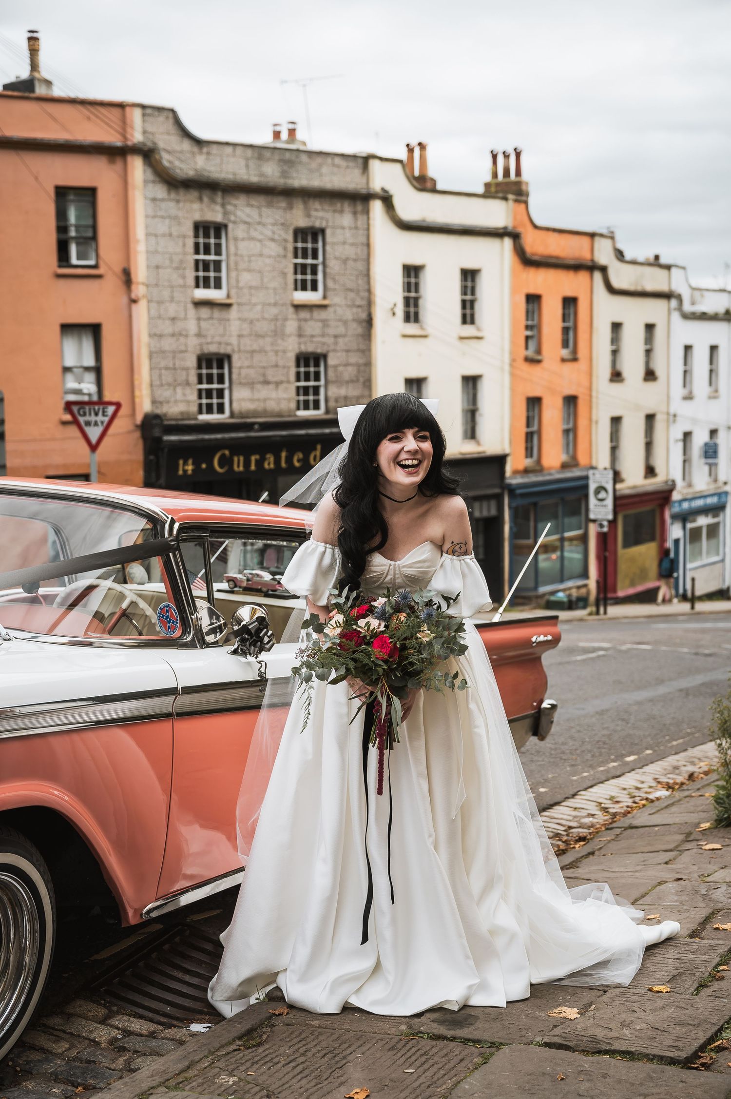 Joyful bride in off-shoulder gown holding bouquet beside vintage pink car on colorful street.