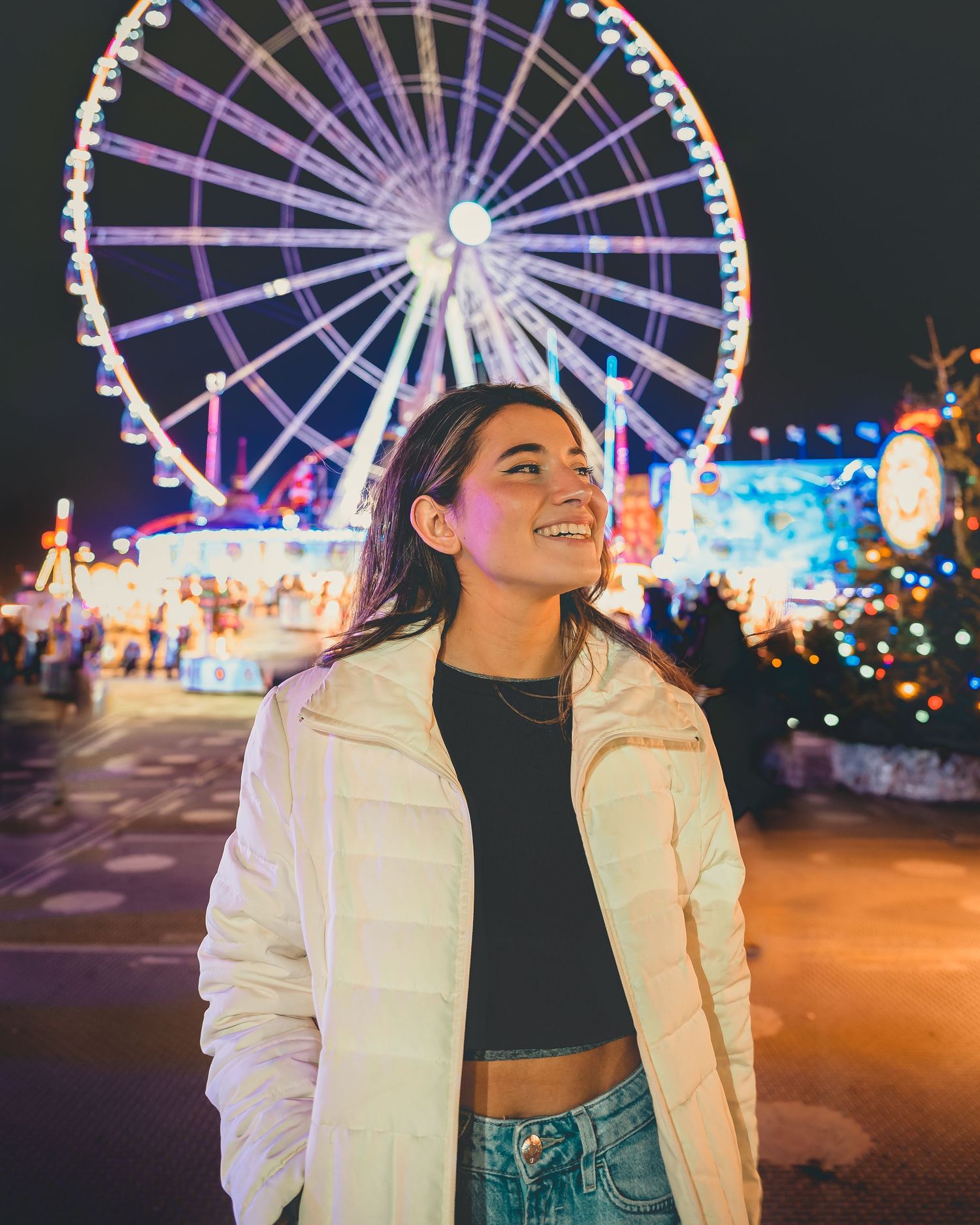 Smiling woman in white puffer jacket and jeans at night carnival with illuminated Ferris wheel behind her.