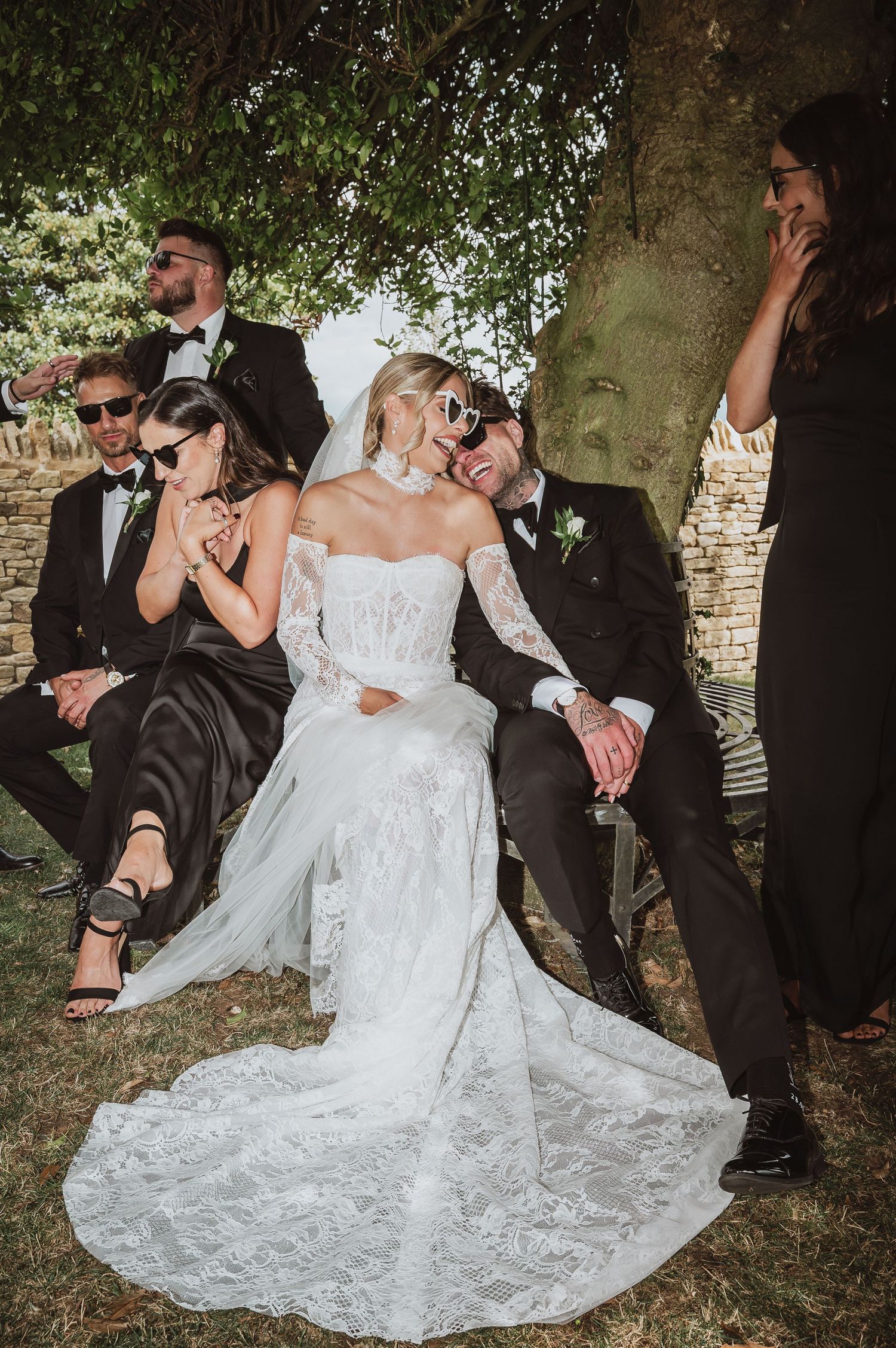 Bride in lace gown and groom in black tuxedo laugh with wedding party under a tree outdoors.