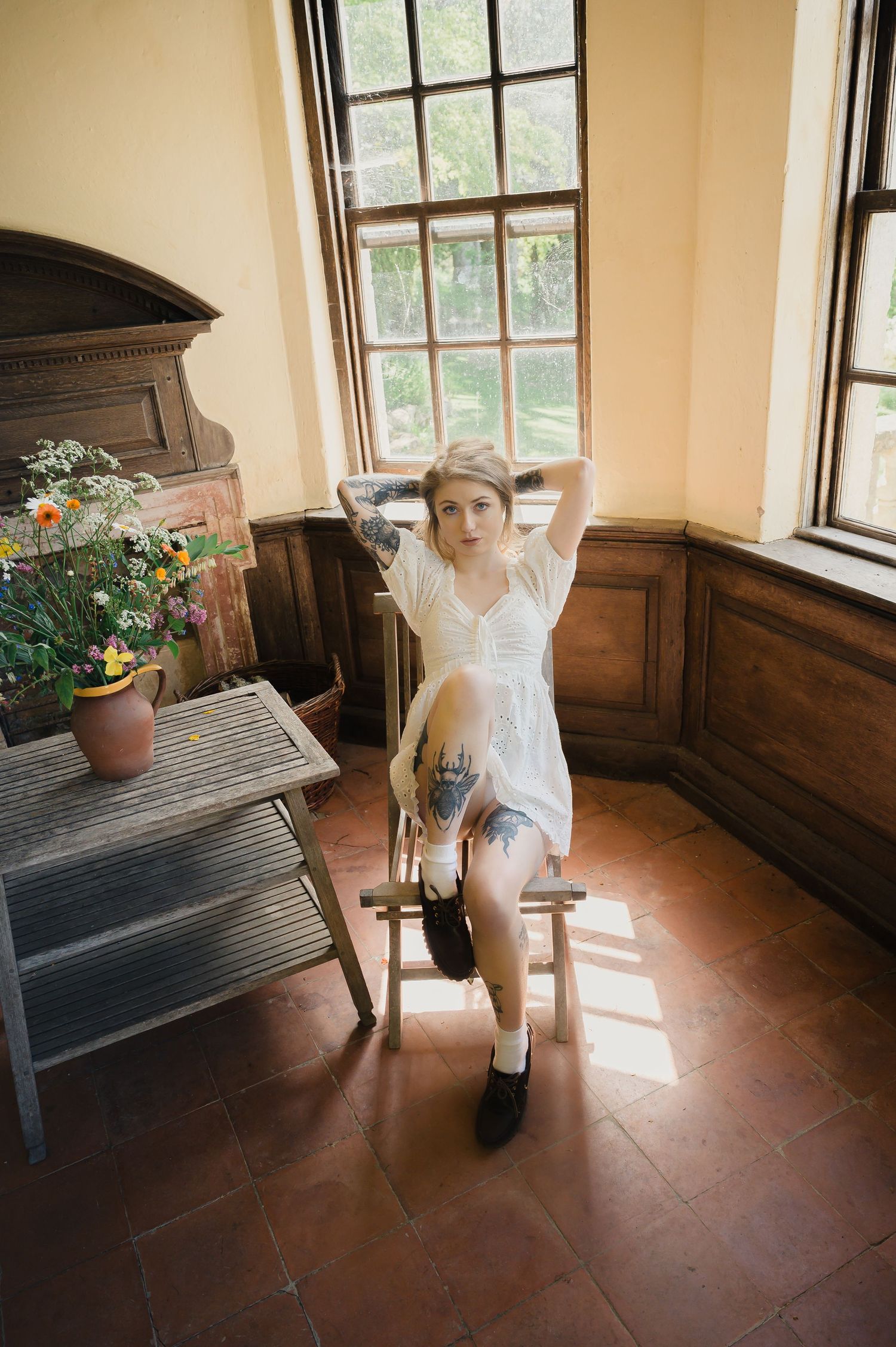 Tattooed woman in white lace dress sits on wooden stool near sunlit windows indoors.