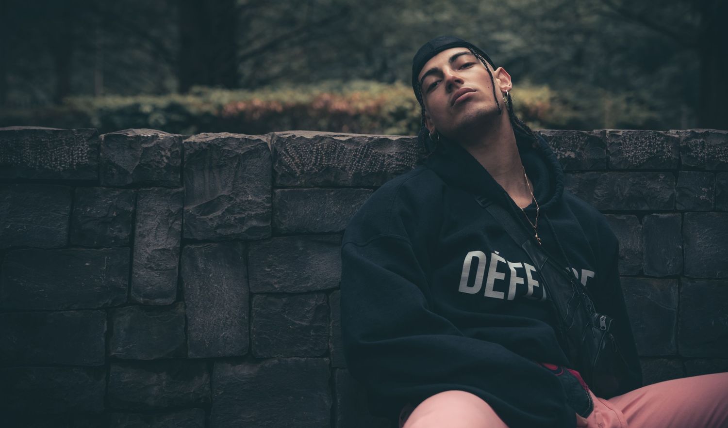 Young man in black hoodie resting against stone wall with eyes closed outdoors.
