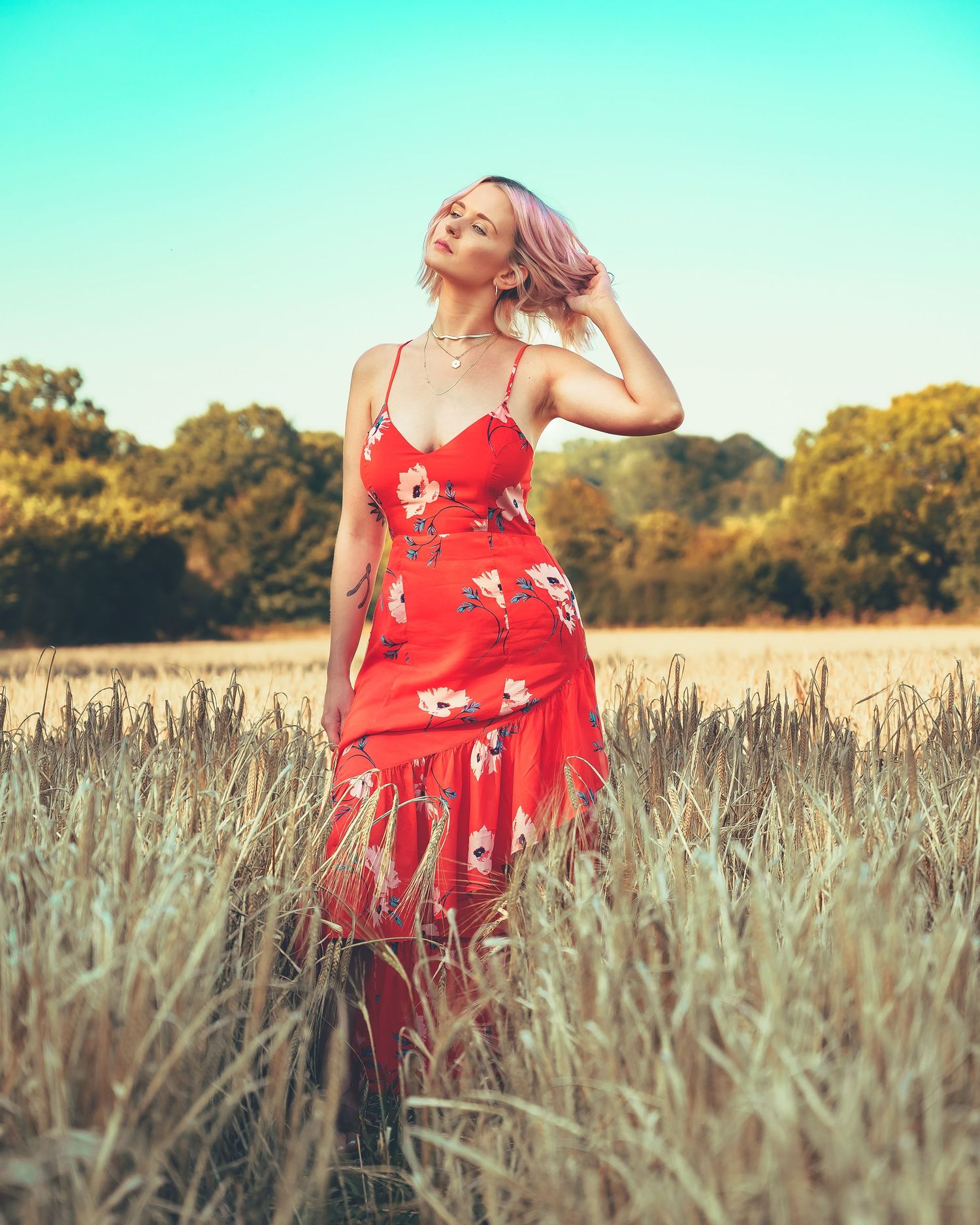 Blonde woman in red floral midi dress stands in a golden wheat field under a clear blue sky.