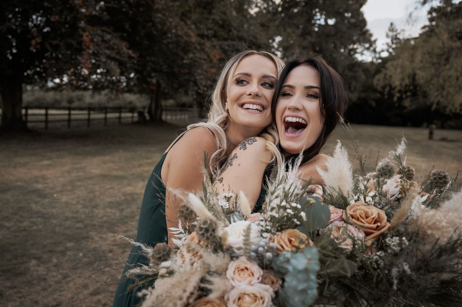 Two bridesmaids in green dresses laughing and embracing while holding wedding bouquets outdoors.