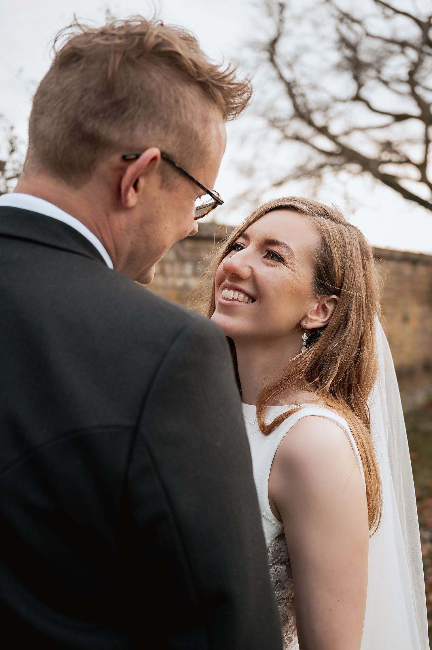 Smiling bride in white veil gazes lovingly at groom wearing glasses during outdoor wedding portraits.