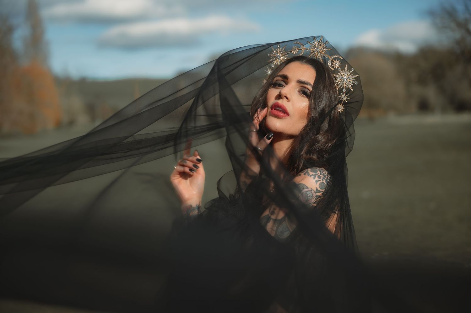 Woman in black lace gown with gold crown and flowing black veil in outdoor setting.