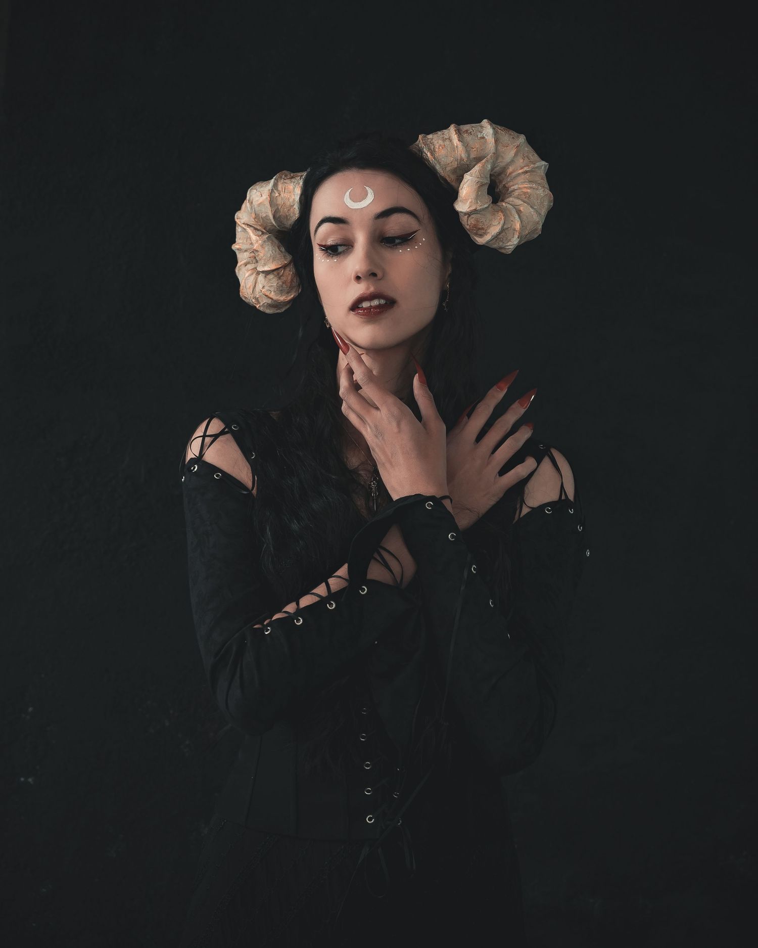 Gothic woman with ram horns and crescent moon makeup posing in black corset against dark background.