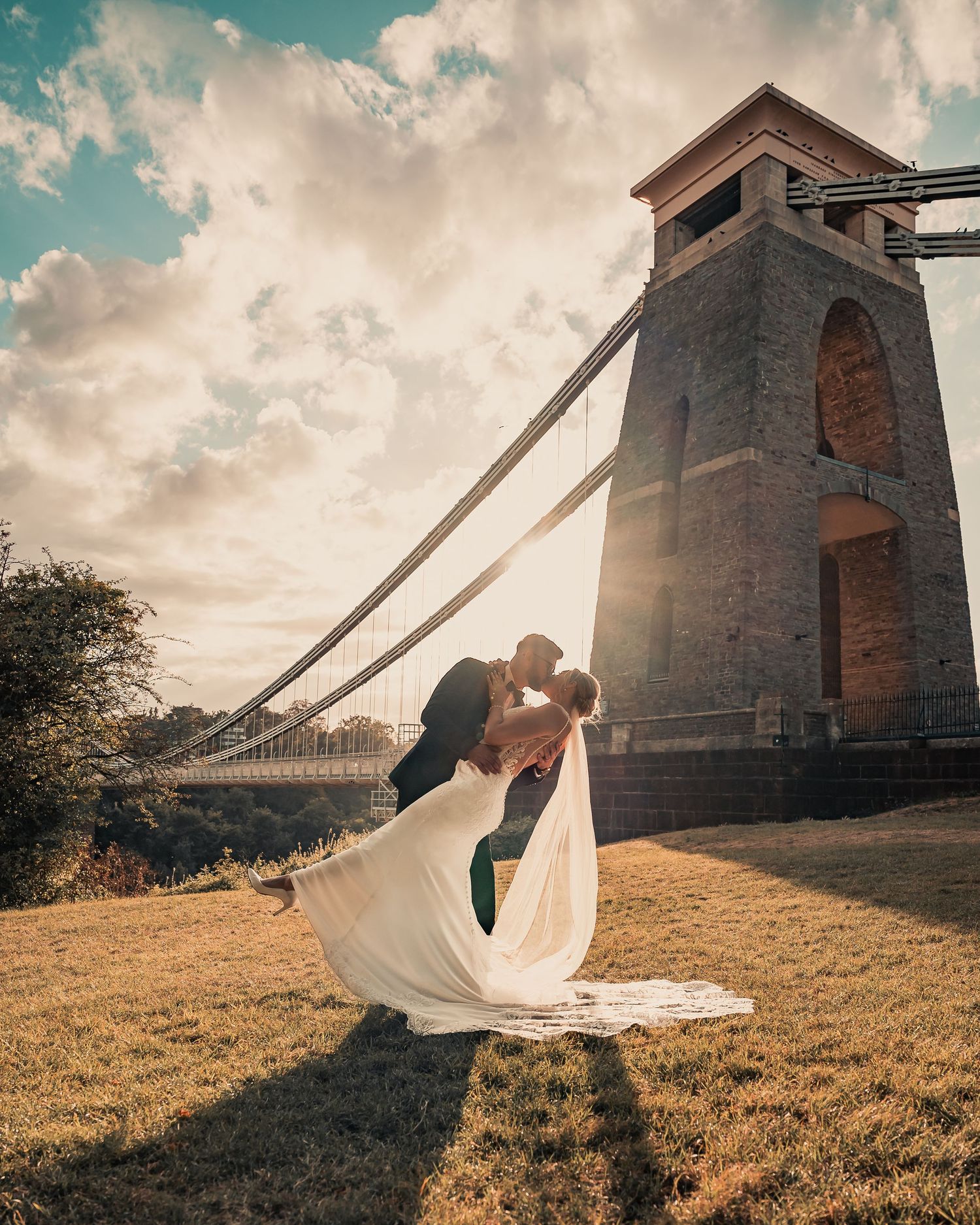 Groom dips bride in a romantic kiss by the Clifton Suspension Bridge tower at sunset.