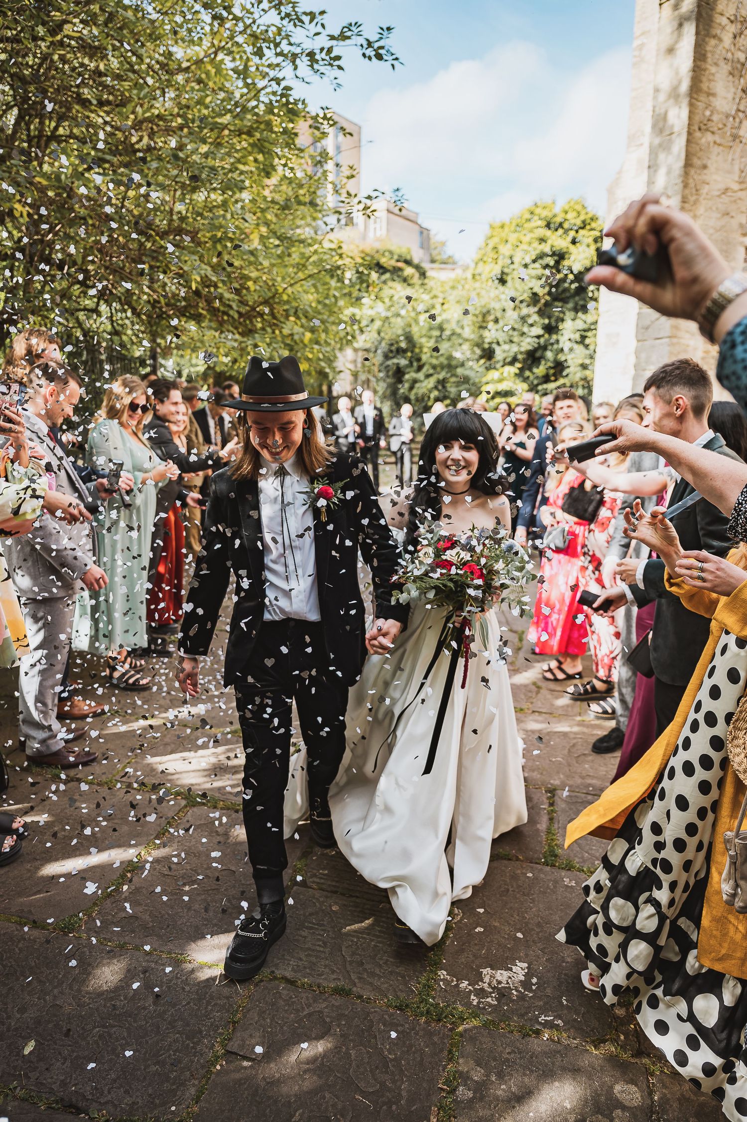 Newlyweds walk through confetti shower with guests cheering outside a church on their wedding day.