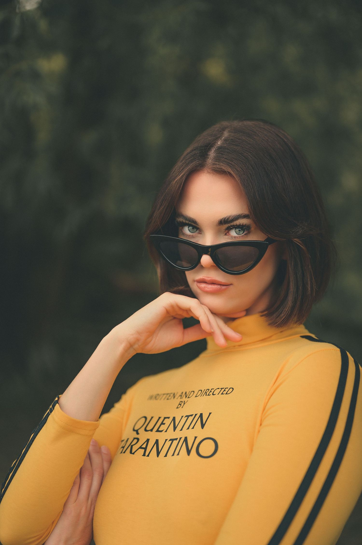 Brunette woman in yellow Quentin Tarantino turtleneck top wearing black cat-eye sunglasses outdoors.
