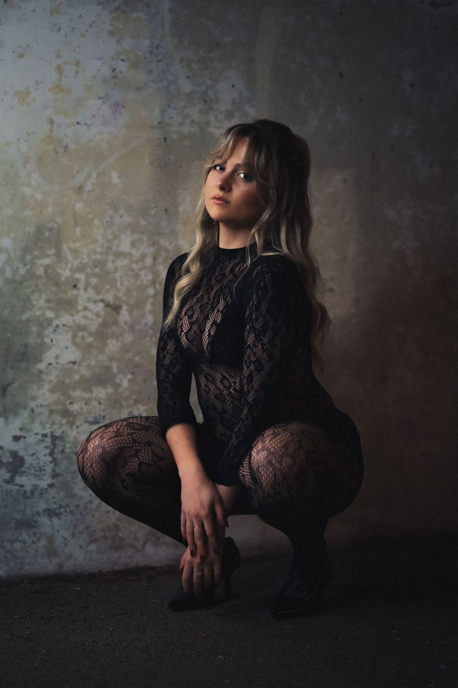 Blonde woman in black lace bodysuit crouching against textured grunge wall in dramatic lighting.
